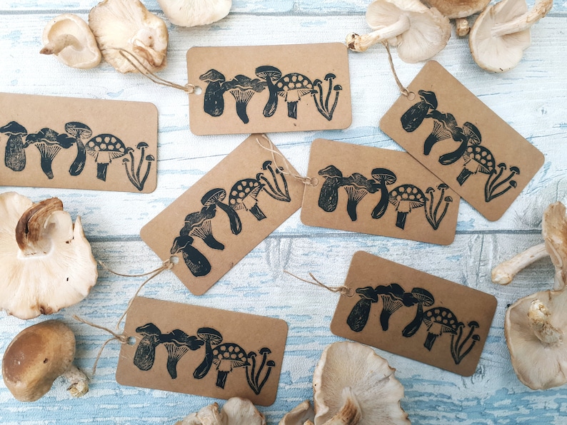 May include: Kraft paper gift tags with a black mushroom design and twine. The tags are rectangular and feature various mushroom illustrations. Several fresh mushrooms are scattered around the tags on a light blue wooden surface.