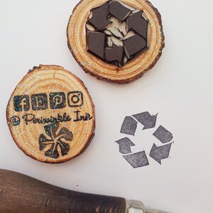 May include: Two wooden stamps with glitter on a white surface. The first stamp has a recycle symbol and the text "@Principle Inn" and social media icons. The second stamp has a recycle symbol. A wooden handle carving tool is also in the image.