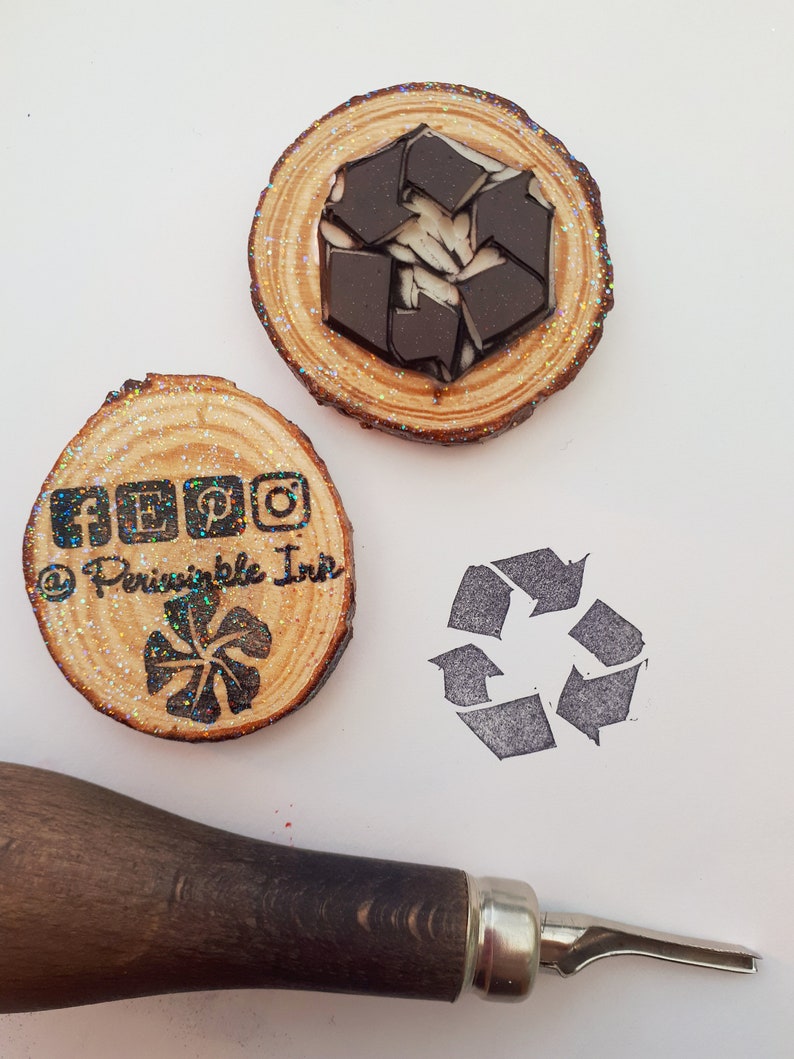May include: Two wooden stamps with glitter on a white surface. The first stamp has a recycle symbol and the text "@Principle Inn" and social media icons. The second stamp has a recycle symbol. A wooden handle carving tool is also in the image.