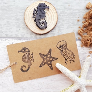 May include: A set of four rubber stamps featuring sea life designs. The stamps are on a brown paper tag with a string attached. The designs include a starfish, a jellyfish, and two seahorses.