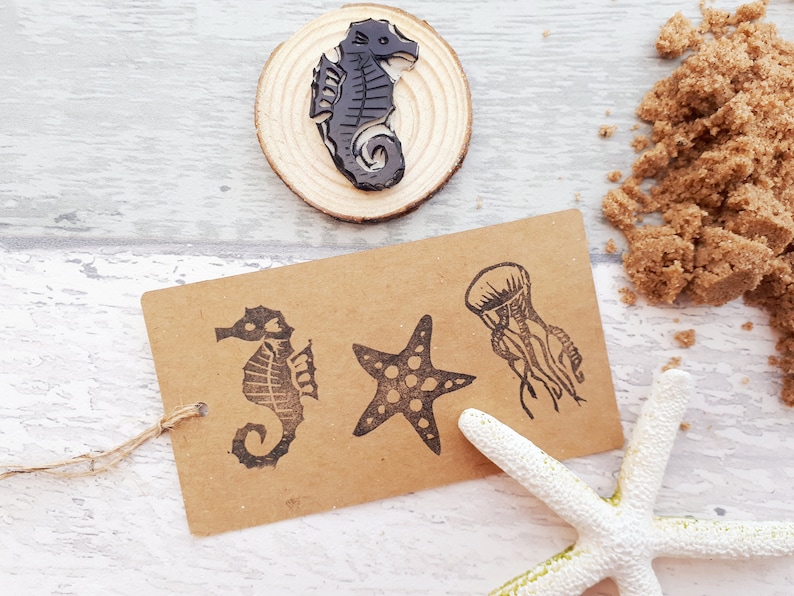 May include: A set of four rubber stamps featuring sea life designs. The stamps are on a brown paper tag with a string attached. The designs include a starfish, a jellyfish, and two seahorses.