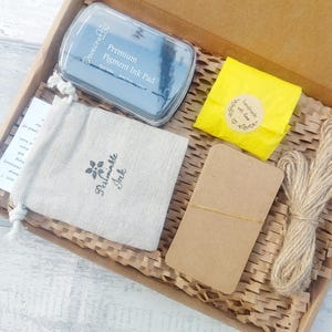May include: A craft box filled with supplies, including a blue ink pad, a drawstring bag with "Periwinkle Ink" printed on it, a yellow package with a sticker, a brown tag, and twine. The box is lined with brown paper.