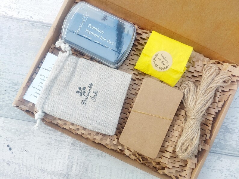 May include: A craft box filled with supplies, including a blue ink pad, a drawstring bag with "Periwinkle Ink" printed on it, a yellow package with a sticker, a brown tag, and twine. The box is lined with brown paper.