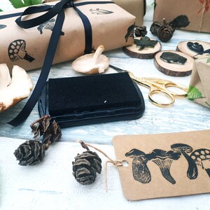 May include: A collection of craft supplies featuring mushroom-themed stamps, ink pad, and gift wrapping. The scene includes brown paper packages tied with black ribbon, wooden slices, and a tag with mushroom illustrations.