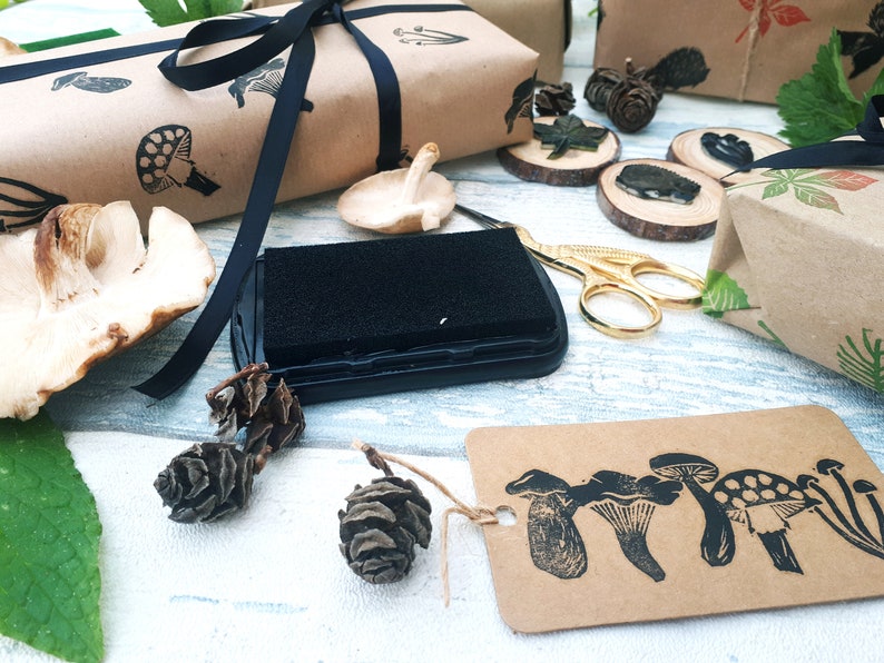 May include: A collection of craft supplies featuring mushroom-themed stamps, ink pad, and gift wrapping. The scene includes brown paper packages tied with black ribbon, wooden slices, and a tag with mushroom illustrations.