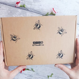 May include: A rectangular cardboard box with five black and white bee illustrations. The box has the social media handles of @Printable Ink. The box is held by two hands, with flowers in the background.
