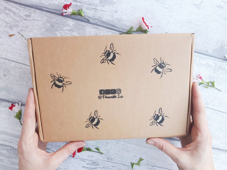 May include: A rectangular cardboard box with five black and white bee illustrations. The box has the social media handles of @Printable Ink. The box is held by two hands, with flowers in the background.