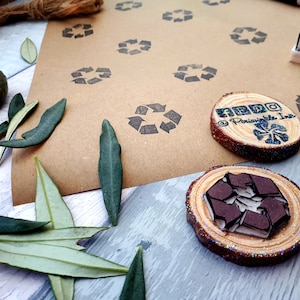 May include: Two wooden stamps with a brown recycled symbol design. The stamps are on a brown paper background with multiple recycled symbol stamps. The stamps are surrounded by green leaves.