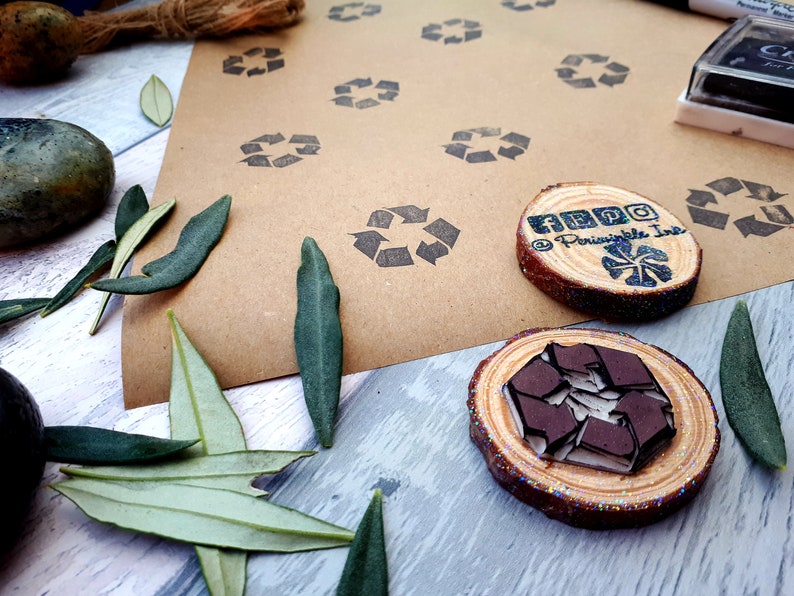 May include: Two wooden stamps with a brown recycled symbol design. The stamps are on a brown paper background with multiple recycled symbol stamps. The stamps are surrounded by green leaves.
