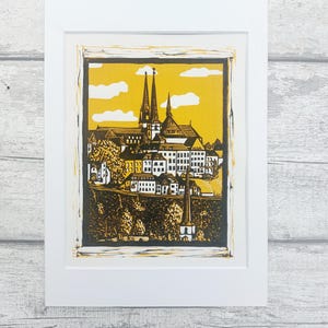 May include: A black and white linocut print of a cityscape with a church in the center. The print is framed in a white mat and a white frame. The background is a yellow color.