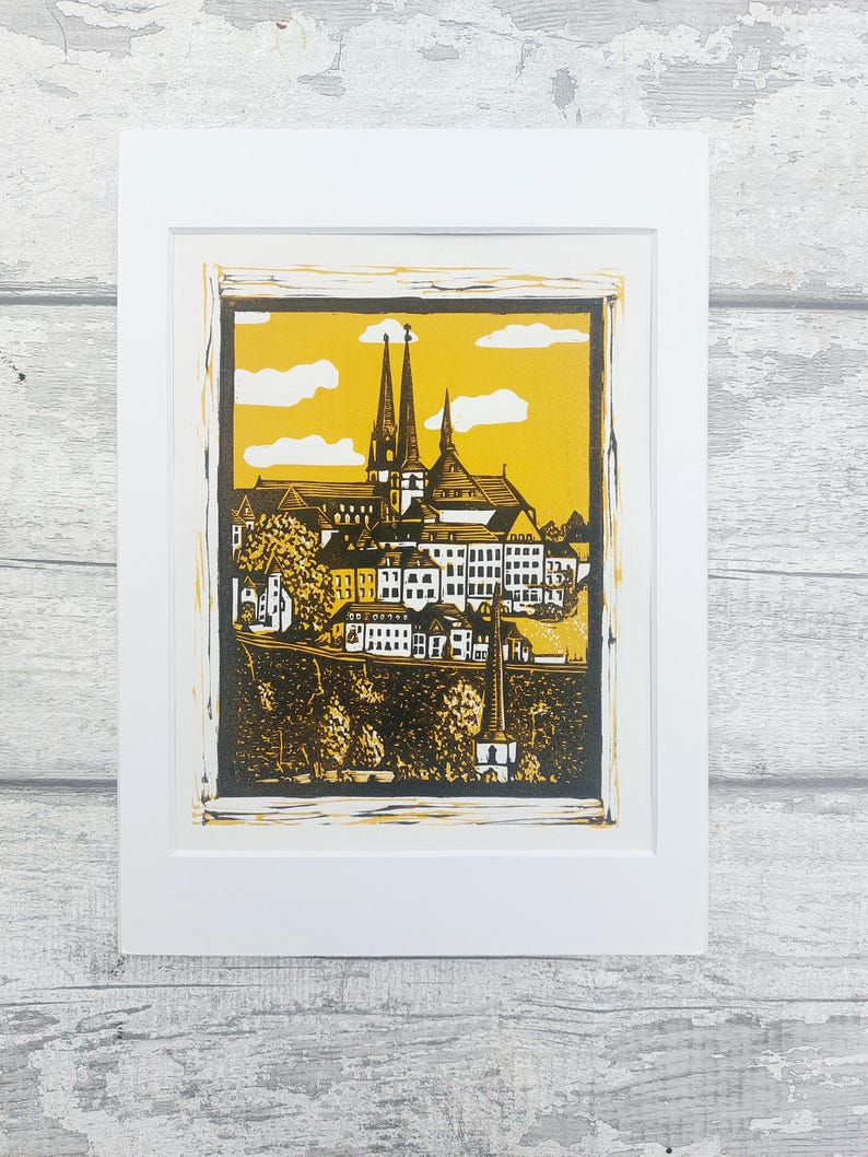 May include: A black and white linocut print of a cityscape with a church in the center. The print is framed in a white mat and a white frame. The background is a yellow color.