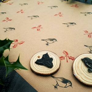 May include: A craft scene featuring a tan paper background with repeating bird and holly leaf stamps in black and red. Two wooden stamps with black designs are in the foreground. Green holly leaves are in the corners.