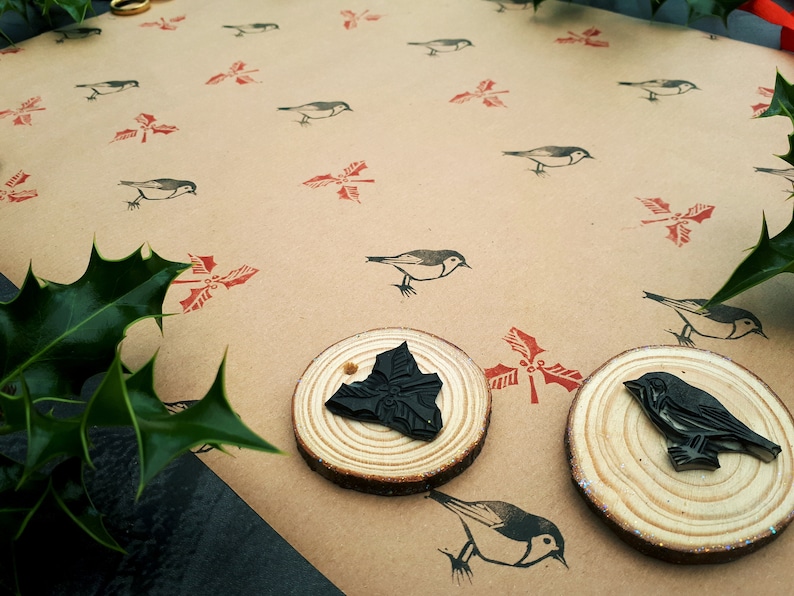 May include: A craft scene featuring a tan paper background with repeating bird and holly leaf stamps in black and red. Two wooden stamps with black designs are in the foreground. Green holly leaves are in the corners.