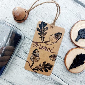 May include: A craft scene featuring a brown tag with acorn and leaf stamps, the word "Merci", and a clear ink pad. Two wood slices with leaf stamps and a real acorn are also present. The ink pad is labeled "Premium Pigment Ink Pad".