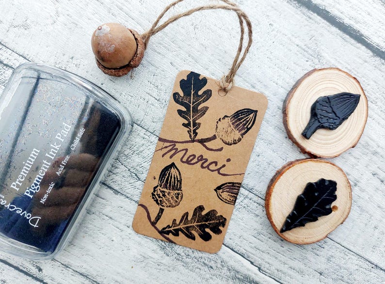 May include: A craft scene featuring a brown tag with acorn and leaf stamps, the word "Merci", and a clear ink pad. Two wood slices with leaf stamps and a real acorn are also present. The ink pad is labeled "Premium Pigment Ink Pad".