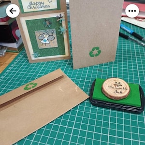 May include: A brown envelope with a green recycle symbol on the front. The envelope is sitting on a green cutting mat. A stamp with a wooden circle and the text "Periwinkle Ink" is in the lower right corner of the image.