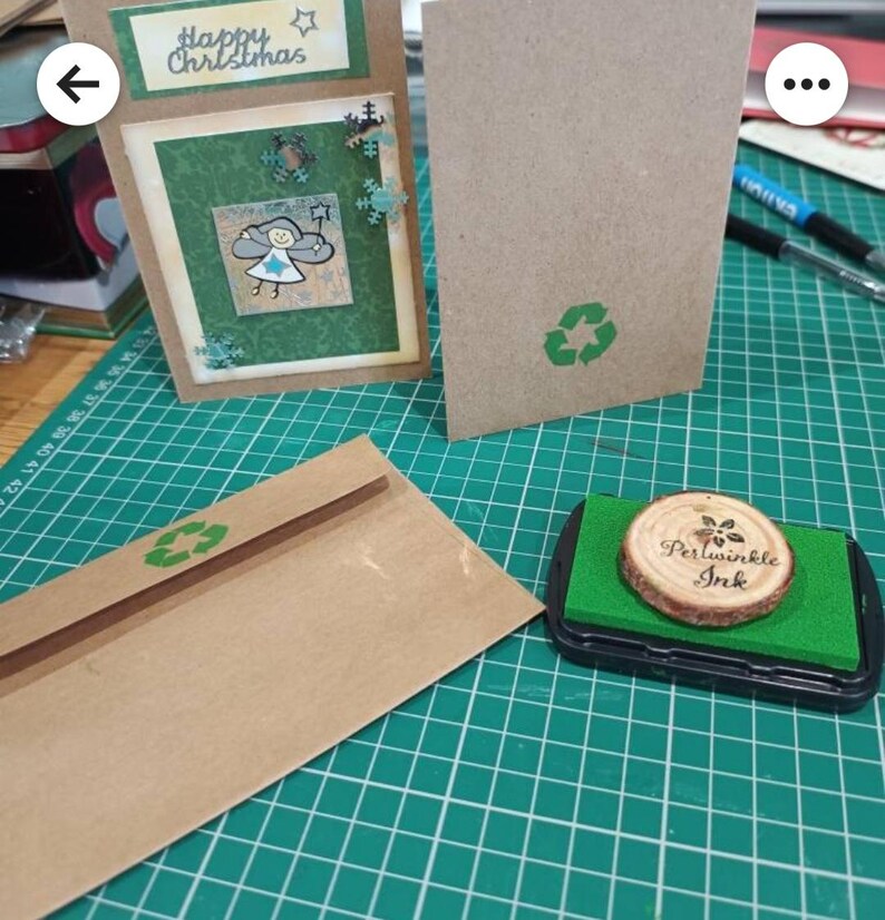 May include: A brown envelope with a green recycle symbol on the front. The envelope is sitting on a green cutting mat. A stamp with a wooden circle and the text "Periwinkle Ink" is in the lower right corner of the image.