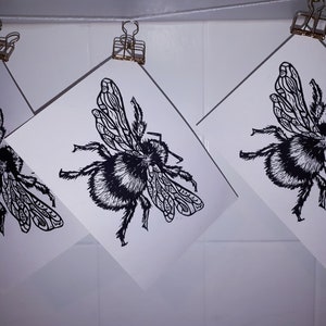 Puede incluir: Tres impresiones rectangulares blancas con una ilustraci&oacute;n en tinta negra de un abejorro con patrones de alas detallados. Cada impresi&oacute;n est&aacute; suspendida de un hilo blanco fino mediante un clip met&aacute;lico dorado, mostrando al insecto desde arriba.