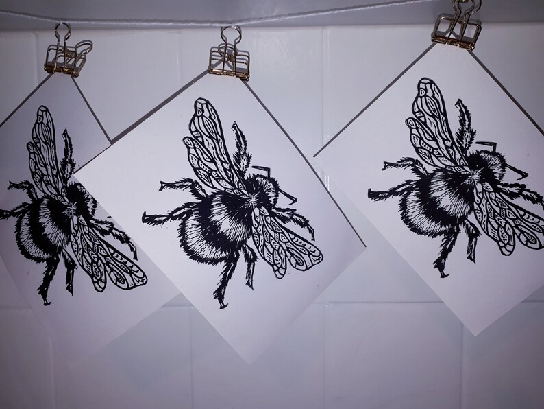 Puede incluir: Tres impresiones rectangulares blancas con una ilustraci&oacute;n en tinta negra de un abejorro con patrones de alas detallados. Cada impresi&oacute;n est&aacute; suspendida de un hilo blanco fino mediante un clip met&aacute;lico dorado, mostrando al insecto desde arriba.