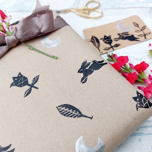 May include: Brown paper gift wrap with a black and silver design featuring a rabbit, leaves, flowers, and a crescent moon. The gift is tied with a brown satin ribbon and decorated with pink flowers.