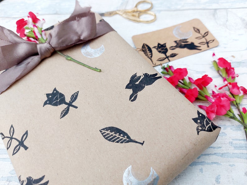 May include: Brown paper gift wrap with a black and silver design featuring a rabbit, leaves, flowers, and a crescent moon. The gift is tied with a brown satin ribbon and decorated with pink flowers.