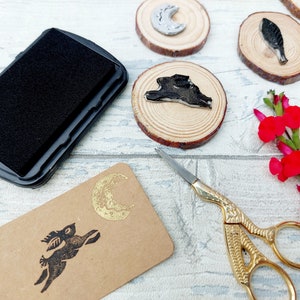 May include: A black ink pad, a gold scissors, and a set of wooden circles with black and gold designs. The designs include a crescent moon, a bird, a rabbit, and a rabbit with a crescent moon.