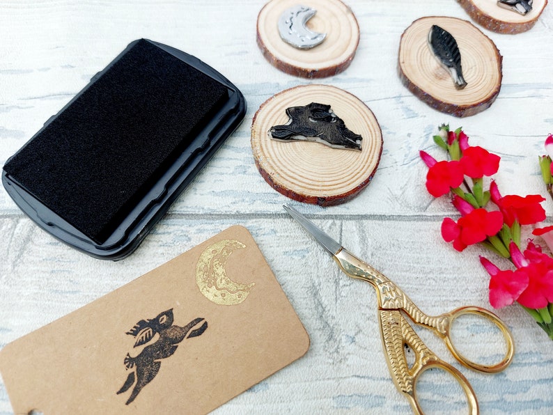 May include: A black ink pad, a gold scissors, and a set of wooden circles with black and gold designs. The designs include a crescent moon, a bird, a rabbit, and a rabbit with a crescent moon.