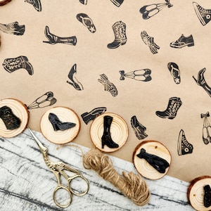 May include: A collection of wooden stamps featuring various shoe designs, including boots, sneakers, and heels. The stamps are arranged on a light brown surface, with a pair of gold scissors and twine nearby. The stamps are on wood slices.