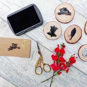 May include: A set of five wooden stamps with black ink designs of a rabbit, a leaf, a flower, a crescent moon, and a moon with a star. The stamps are on a white wooden surface with a black ink pad, a pair of gold scissors, and a brown tag with a black ink design of a rabbit and a gold crescent moon.