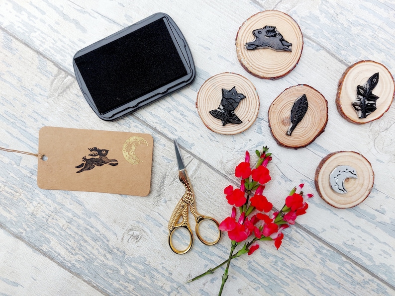 May include: A set of five wooden stamps with black ink designs of a rabbit, a leaf, a flower, a crescent moon, and a moon with a star. The stamps are on a white wooden surface with a black ink pad, a pair of gold scissors, and a brown tag with a black ink design of a rabbit and a gold crescent moon.