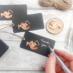 May include: Black gift tags with a copper-colored illustration of a fish with a large mouth. The tags are tied with twine and have the words "Thank you!" written on one of them.