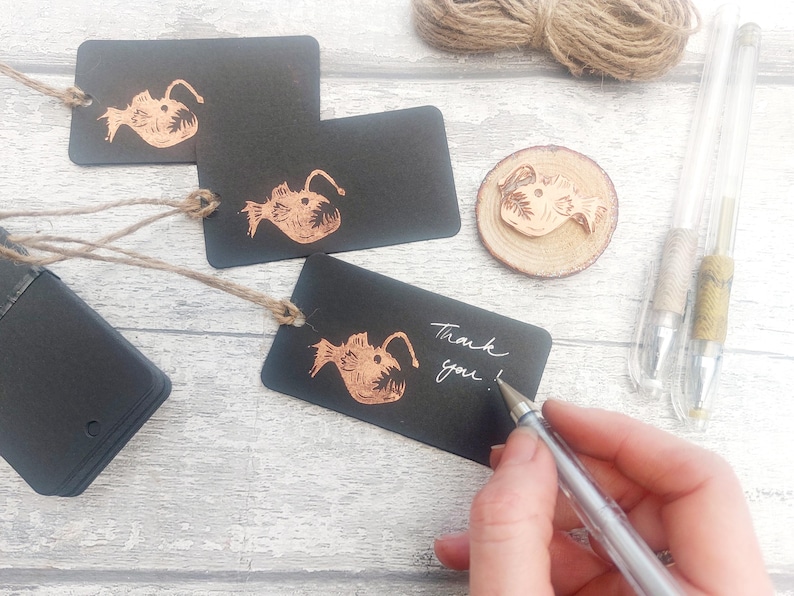 May include: Black gift tags with a copper-colored illustration of a fish with a large mouth. The tags are tied with twine and have the words "Thank you!" written on one of them.