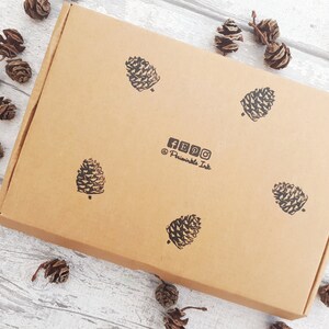 May include: A brown cardboard box with black pine cone stamps. The box has the social media icons for Facebook, Instagram, and Pinterest, along with the text "@Printable Ink". Small pine cones are scattered around the box.