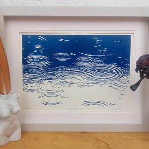 May include: A framed print depicting a blue and white abstract image of raindrops creating ripples on water. The artwork is in a white frame. A decorative feather and a bust sculpture are on the left, and a dark flower in a vase is on the right.
