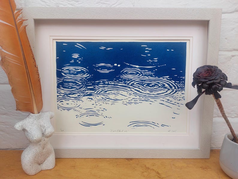 May include: A framed print depicting a blue and white abstract image of raindrops creating ripples on water. The artwork is in a white frame. A decorative feather and a bust sculpture are on the left, and a dark flower in a vase is on the right.