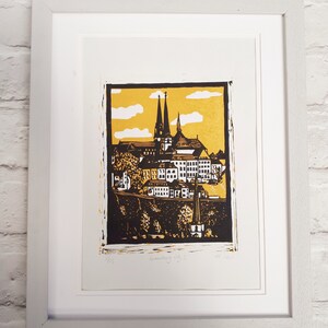 May include: A framed black and white linocut print of a cityscape with two tall church spires. The print has a yellow background with white clouds and the text "Luxembourg City I" and "2021" in the bottom right corner.
