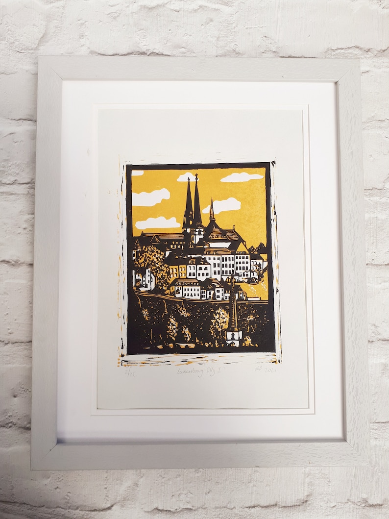 May include: A framed black and white linocut print of a cityscape with two tall church spires. The print has a yellow background with white clouds and the text "Luxembourg City I" and "2021" in the bottom right corner.