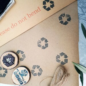 May include: A brown paper package with a stamp that says "Please do not bend". The package is decorated with multiple black recycle symbols and a brown twine bow. There are two wooden circles with a black recycle symbol and a blue and white logo.