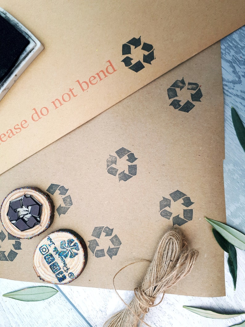 May include: A brown paper package with a stamp that says "Please do not bend". The package is decorated with multiple black recycle symbols and a brown twine bow. There are two wooden circles with a black recycle symbol and a blue and white logo.