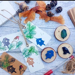 May include: A collection of handmade gift tags with birdhouse and bird illustrations, along with wooden slices, twine, and natural elements like pinecones and seed pods. The tags are white and brown with green and blue accents.