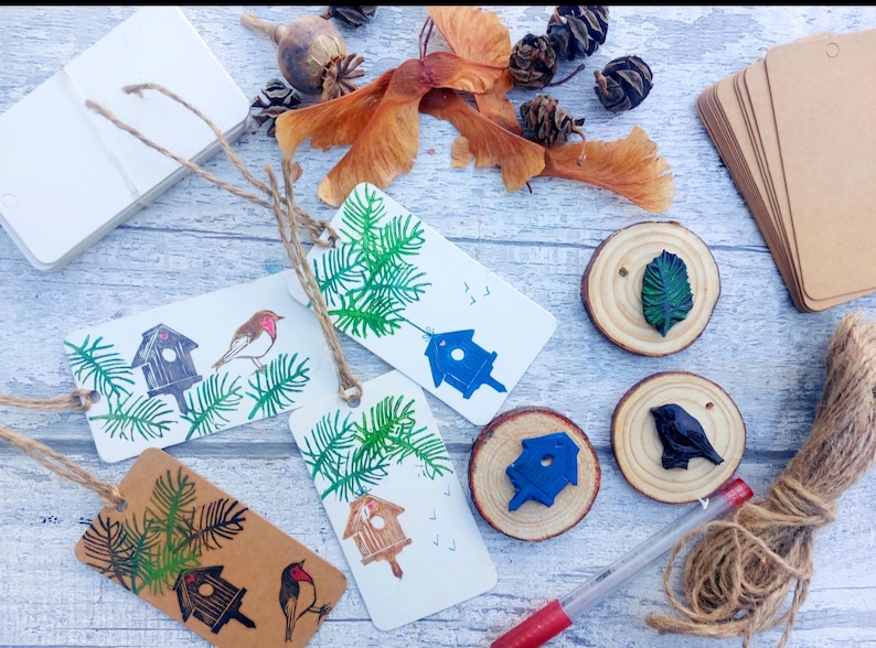 May include: A collection of handmade gift tags with birdhouse and bird illustrations, along with wooden slices, twine, and natural elements like pinecones and seed pods. The tags are white and brown with green and blue accents.