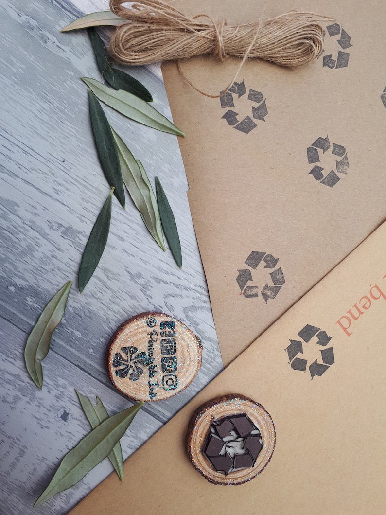 May include: Two wooden round slices with a brown background. One slice has a black and white recycle symbol and the other has a black and white swirl design. Both slices have a glittery blue finish. The background is brown paper with a recycle symbol printed on it.