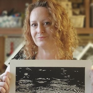 May include: A black and white linocut print depicting raindrops falling on water. The artwork is held by a person with curly red hair. The print shows a close-up of the water's surface with concentric circles.