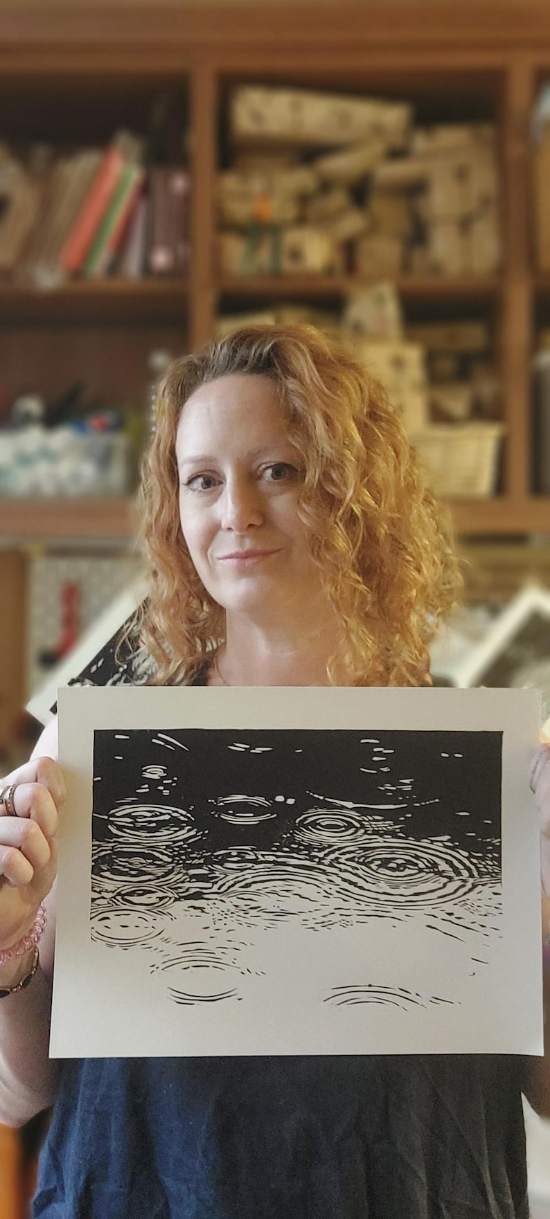 May include: A black and white linocut print depicting raindrops falling on water. The artwork is held by a person with curly red hair. The print shows a close-up of the water's surface with concentric circles.