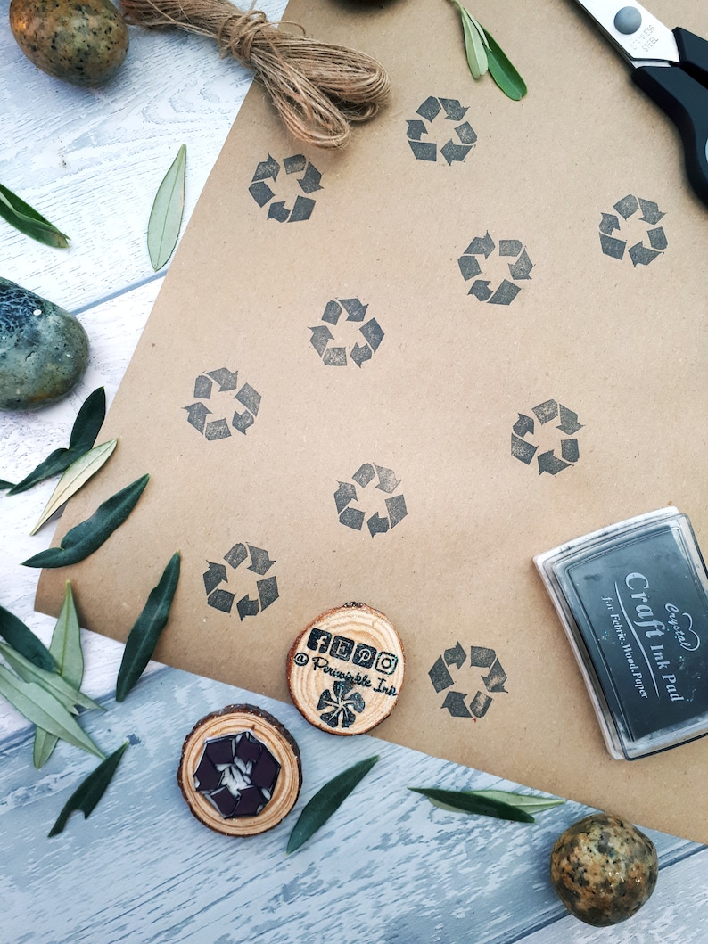 May include: A brown paper sheet with multiple black recycle symbols stamped on it. There is a wooden stamp with the text "Periwinkle Ink" and a black ink pad with the text "Crystal Craft Ink Pad for Fabric, Wood, Paper".