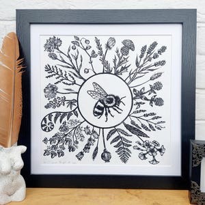May include: Framed black and white art print depicting a bee encircled by botanical illustrations. The print is in a black frame, with a feather and a small statue nearby. The artwork is set against a white background. The print measures approximately 30 cm x 30 cm.