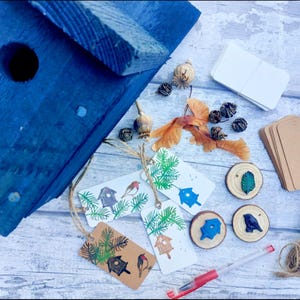 May include: A blue wooden birdhouse with a round entry hole is surrounded by craft supplies. Includes tags with birdhouse and bird illustrations, wooden slices, twine, and blank tags. The items are arranged on a light wood-grain surface.