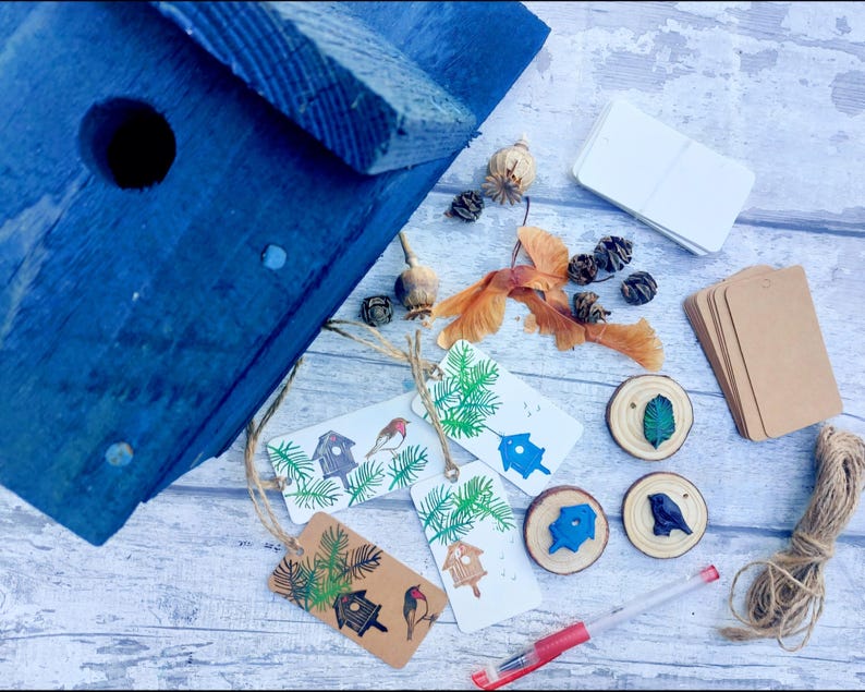 May include: A blue wooden birdhouse with a round entry hole is surrounded by craft supplies. Includes tags with birdhouse and bird illustrations, wooden slices, twine, and blank tags. The items are arranged on a light wood-grain surface.