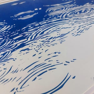 May include: Blue and white linocut print depicting a water ripple effect. The artwork features concentric circles and curved lines, creating a sense of movement. The title "Ripple Effect" is handwritten in the lower right corner.