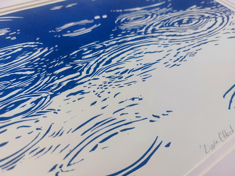 May include: Blue and white linocut print depicting a water ripple effect. The artwork features concentric circles and curved lines, creating a sense of movement. The title "Ripple Effect" is handwritten in the lower right corner.